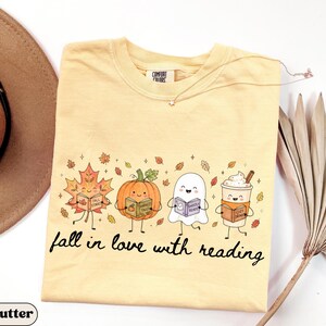 Peut inclure: Un t-shirt jaune beurre avec un motif repr&eacute;sentant une feuille d'&eacute;rable, une citrouille, un fant&ocirc;me et une tasse de caf&eacute; souriants, chacun tenant un livre. Le texte en dessous indique fall in love with reading. &Eacute;tiquette Comfort Colors au col.
