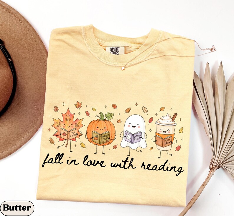 Peut inclure: Un t-shirt jaune beurre avec un motif repr&eacute;sentant une feuille d'&eacute;rable, une citrouille, un fant&ocirc;me et une tasse de caf&eacute; souriants, chacun tenant un livre. Le texte en dessous indique fall in love with reading. &Eacute;tiquette Comfort Colors au col.