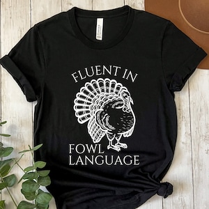 May include: Black t-shirt featuring a white turkey illustration and the text "FLUENT IN FOWL LANGUAGE". The turkey is depicted with a fanned tail. The shirt is made of a soft material and is suitable for casual wear.