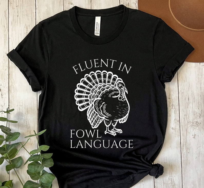May include: Black t-shirt featuring a white turkey illustration and the text "FLUENT IN FOWL LANGUAGE". The turkey is depicted with a fanned tail. The shirt is made of a soft material and is suitable for casual wear.