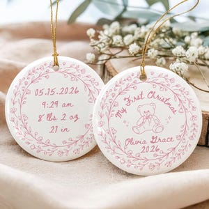 May include: Two round white ceramic ornaments with gold hanging strings. One features a pink floral wreath, the text 05.15.2026, 9:29 am, 8 lbs 2 oz, and 21 in. The other shows a pink floral wreath, a teddy bear, My First Christmas, Olivia Grace, and 2026.