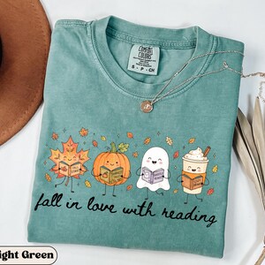 Peut inclure: Un t-shirt &agrave; manches courtes vert clair avec un motif repr&eacute;sentant une feuille d'&eacute;rable, une citrouille, un fant&ocirc;me et une tasse de caf&eacute;, chacun tenant un livre. Le texte "fall in love with reading" est imprim&eacute; sous les personnages.