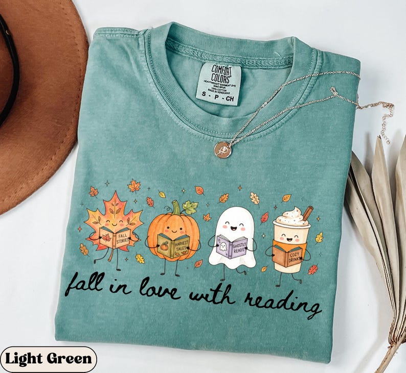 Peut inclure: Un t-shirt &agrave; manches courtes vert clair avec un motif repr&eacute;sentant une feuille d'&eacute;rable, une citrouille, un fant&ocirc;me et une tasse de caf&eacute;, chacun tenant un livre. Le texte "fall in love with reading" est imprim&eacute; sous les personnages.