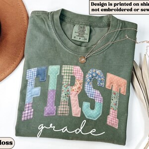 May include: A moss green t-shirt with the word "FIRST" in patchwork letters and the word "grade" in script. The patchwork letters are in various colors and patterns. A gold necklace with a pendant is on the shirt.