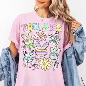 May include: Pink t-shirt with the text "You Are" in rainbow colors. The shirt features colorful bunny illustrations with words like "Capable", "Worthy", "Enough", "Strong", and "Loved".