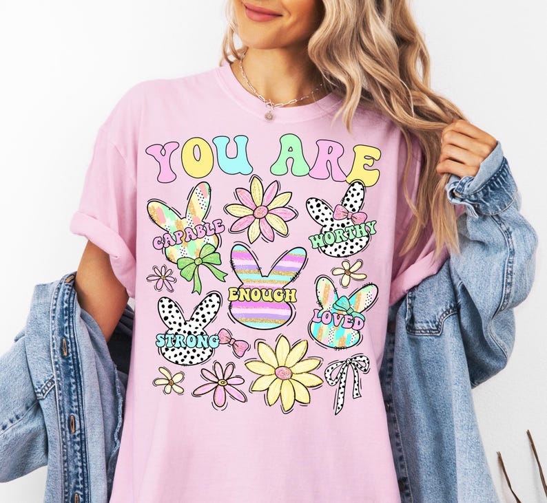 May include: Pink t-shirt with the text "You Are" in rainbow colors. The shirt features colorful bunny illustrations with words like "Capable", "Worthy", "Enough", "Strong", and "Loved".
