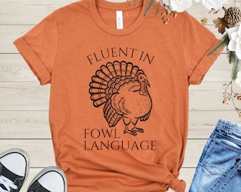May include: An orange t-shirt with the text "FLUENT IN FOWL LANGUAGE" above a black illustration of a turkey. The shirt is folded at the bottom and paired with blue jeans and black sneakers.