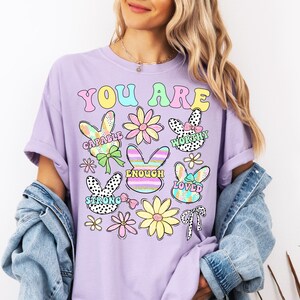 May include: A lavender t-shirt with the text "You Are" in rainbow colors. The shirt features a variety of colorful bunny and flower illustrations with positive affirmations like "Capable", "Worthy", "Enough", "Loved", and "Strong".