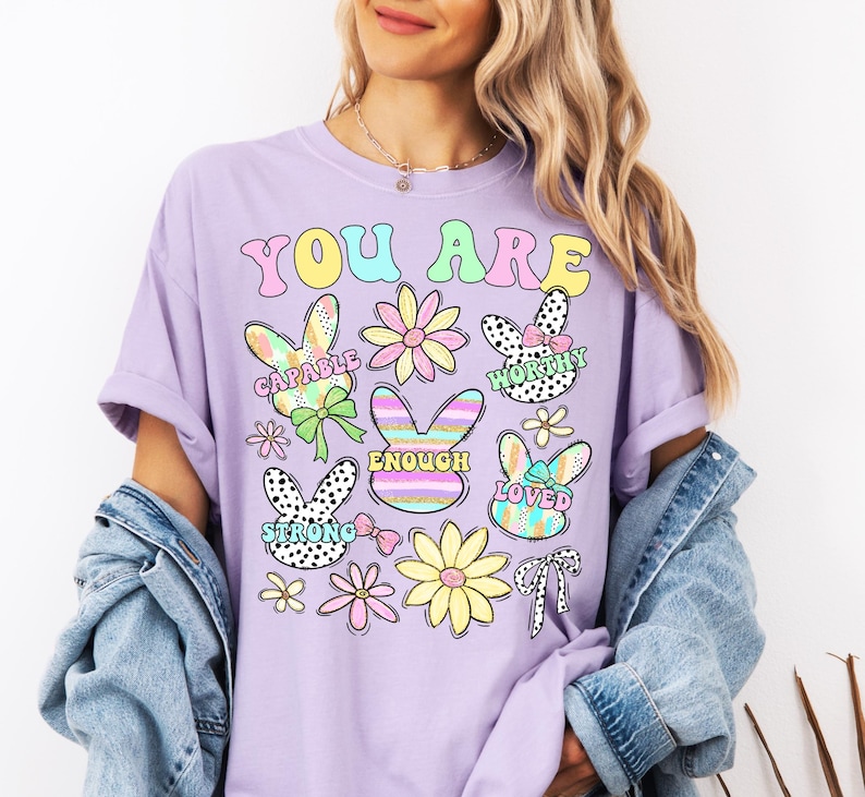 May include: A lavender t-shirt with the text "You Are" in rainbow colors. The shirt features a variety of colorful bunny and flower illustrations with positive affirmations like "Capable", "Worthy", "Enough", "Loved", and "Strong".