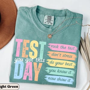 May include: Light green t-shirt with the words "TEST DAY" in colorful block letters. The shirt also has the phrases "you got this", "rock the test", "don't stress", "do your best", "you know it", and "now show it". A silver necklace with a pendant is on the shirt.