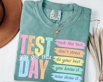 May include: Light green t-shirt with the words "TEST DAY" in colorful block letters. The shirt also has the phrases "you got this", "rock the test", "don't stress", "do your best", "you know it", and "now show it". A silver necklace with a pendant is on the shirt.