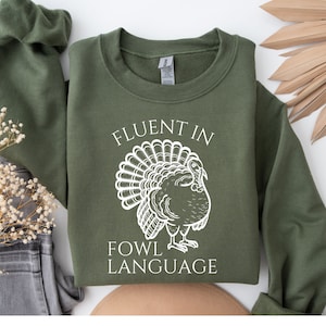 May include: An olive green sweatshirt with the text "FLUENT IN FOWL LANGUAGE" and a white turkey graphic. The sweatshirt is folded, with a matching sleeve visible. The design is centered on the front.
