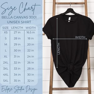 Pu&ograve; includere: Tabella delle taglie per magliette unisex Bella Canvas 3001. La tabella mostra la lunghezza e la larghezza in pollici per le taglie XS a 5XL. Una maglietta nera &egrave; mostrata su una scala di legno.
