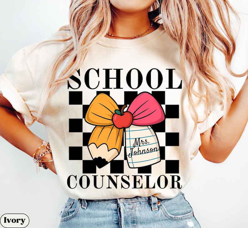 May include: Ivory-colored t-shirt with the words "SCHOOL COUNSELOR" in black block letters. The design features a cartoon pencil and pink bow with an apple and a notepad that says "Mrs. Johnson".