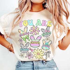 May include: A cream-colored t-shirt with the text "YOU ARE" in rainbow colors. The shirt features colorful bunny illustrations with positive affirmations like "CAPABLE", "WORTHY", "ENOUGH", "LOVED", and "STRONG".