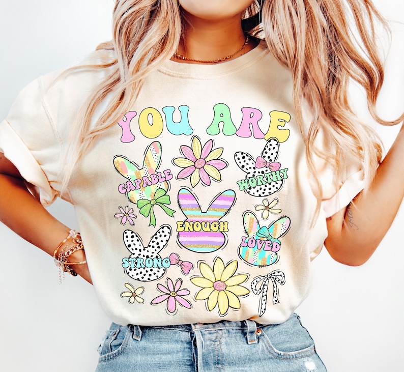 May include: A cream-colored t-shirt with the text "YOU ARE" in rainbow colors. The shirt features colorful bunny illustrations with positive affirmations like "CAPABLE", "WORTHY", "ENOUGH", "LOVED", and "STRONG".