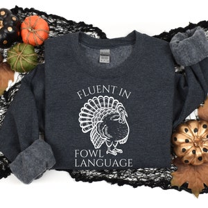 May include: Dark gray sweatshirt with the text "FLUENT IN FOWL LANGUAGE" and a white turkey illustration. The sweatshirt is surrounded by fall-themed decorations, including pumpkins and leaves.