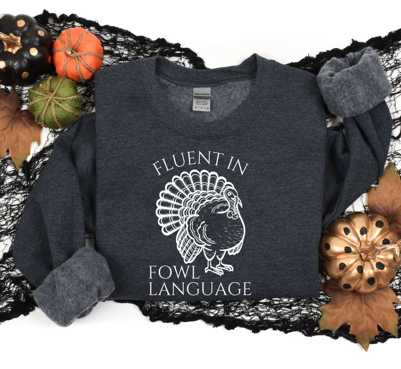 May include: Dark gray sweatshirt with the text "FLUENT IN FOWL LANGUAGE" and a white turkey illustration. The sweatshirt is surrounded by fall-themed decorations, including pumpkins and leaves.
