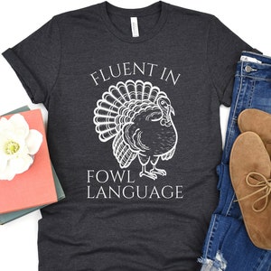 May include: Dark grey t-shirt featuring a white turkey illustration and the text "FLUENT IN FOWL LANGUAGE". The shirt is folded, with blue jeans and brown shoes visible. A book and a white flower are also in the image.