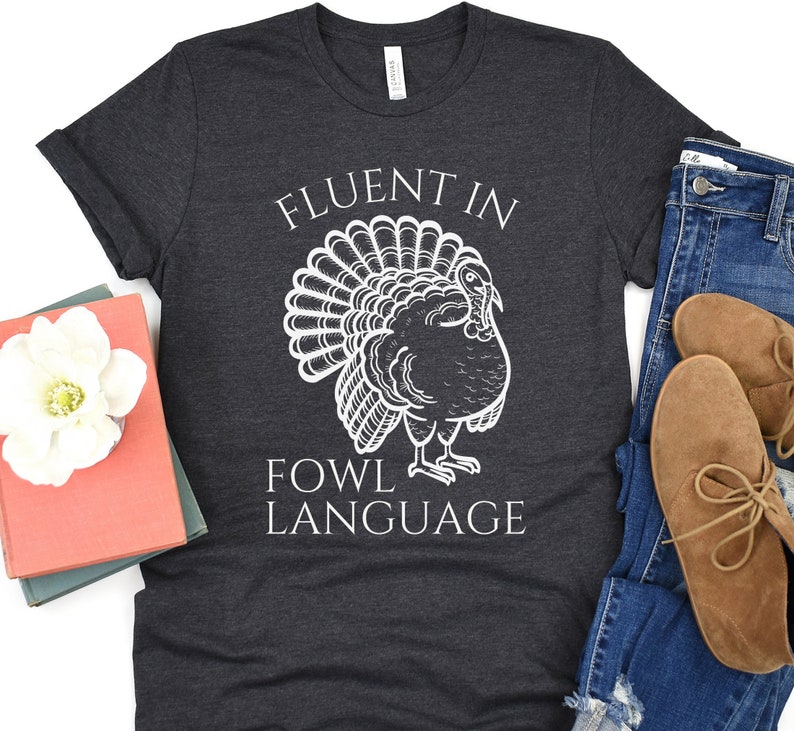 May include: Dark grey t-shirt featuring a white turkey illustration and the text "FLUENT IN FOWL LANGUAGE". The shirt is folded, with blue jeans and brown shoes visible. A book and a white flower are also in the image.