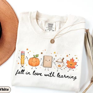 May include: A cream-colored t-shirt featuring a graphic of a smiling pencil, pumpkin, notebook, coffee mug, and maple leaf. The text "fall in love with learning" is printed in black script below the illustrations, surrounded by small gold stars.
