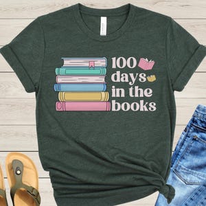 Pu&ograve; includere: Una maglietta verde scuro con una pila di libri colorati e il testo "100 giorni tra i libri".