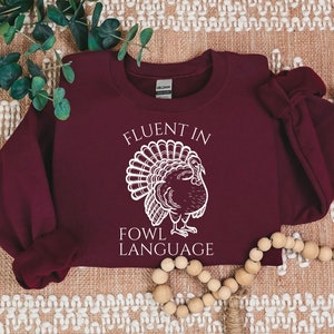 May include: A maroon sweatshirt with the text "FLUENT IN FOWL LANGUAGE" and a white illustration of a turkey. The sweatshirt is a crew neck style, perfect for fall and Thanksgiving.
