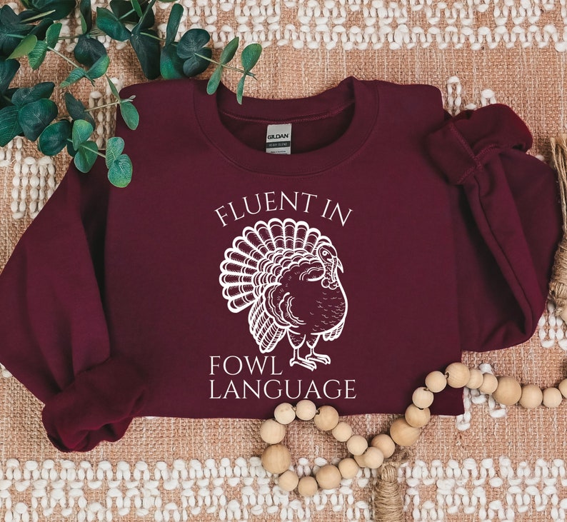 May include: A maroon sweatshirt with the text "FLUENT IN FOWL LANGUAGE" and a white illustration of a turkey. The sweatshirt is a crew neck style, perfect for fall and Thanksgiving.