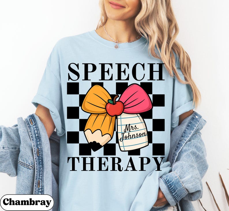 May include: Light blue t-shirt with the words "SPEECH THERAPY" in black block letters. The design includes a pencil, a pink bow, and a red apple. A notepad with the name "Mrs. Johnson" is also featured.