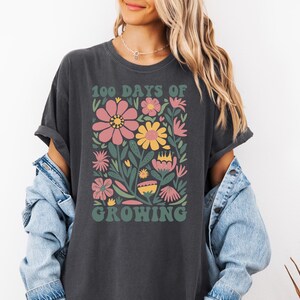 May include: A dark gray t-shirt with a floral design and the text "100 Days of Growing" in a retro font.