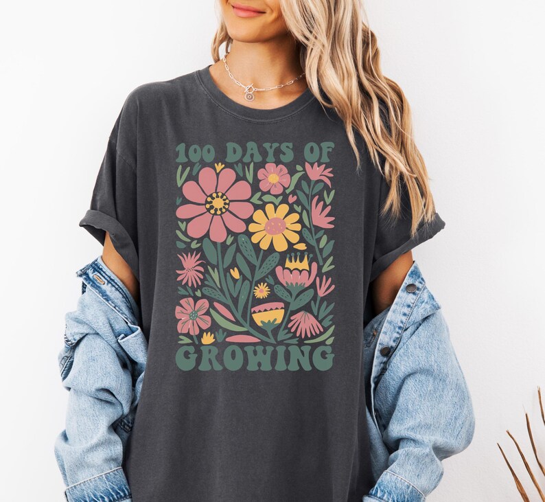 May include: A dark gray t-shirt with a floral design and the text "100 Days of Growing" in a retro font.