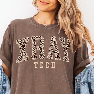 May include: A brown t-shirt with the words "XRAY TECH" printed in white letters. The word "XRAY" is in a leopard print font.