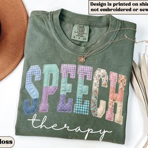 May include: A moss green t-shirt with the word "SPEECH" in patchwork letters and "therapy" in cursive. The patchwork letters are in various patterns and colors. The shirt has the text "Design is printed on shirt, not embroidered or sewn".