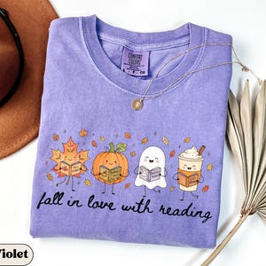 Peut inclure: Un t-shirt violet &agrave; manches courtes avec une impression graphique repr&eacute;sentant une feuille d'&eacute;rable, une citrouille, un fant&ocirc;me et une tasse de caf&eacute; souriants, chacun tenant un livre. Le texte "fall in love with reading" est imprim&eacute; en dessous.