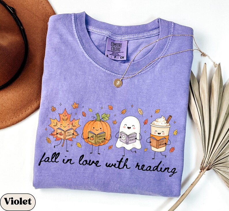 Peut inclure: Un t-shirt violet &agrave; manches courtes avec une impression graphique repr&eacute;sentant une feuille d'&eacute;rable, une citrouille, un fant&ocirc;me et une tasse de caf&eacute; souriants, chacun tenant un livre. Le texte "fall in love with reading" est imprim&eacute; en dessous.