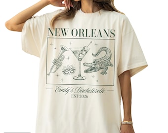 May include: Ivory-colored t-shirt with a moss green graphic featuring a trumpet, martini, mask, and alligator, with the text "NEW ORLEANS" and "Emily's Bachelorette EST 2026".