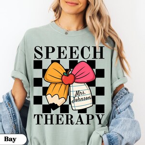 May include: A light green t-shirt with the words "SPEECH THERAPY" in black. The shirt features a graphic of a pencil, bow, and apple on a checkered background. A small notepad reads "Mrs. Johnson."