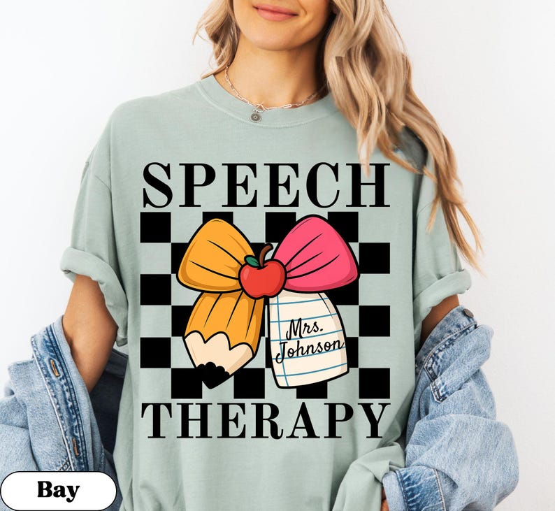May include: A light green t-shirt with the words "SPEECH THERAPY" in black. The shirt features a graphic of a pencil, bow, and apple on a checkered background. A small notepad reads "Mrs. Johnson."