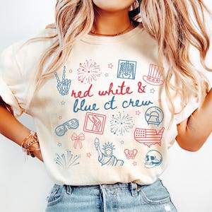 May include: Cream-colored t-shirt with the text "Red White & Blue CT Crew" and various red and blue graphic illustrations. The illustrations include fireworks, the Statue of Liberty, and a skeleton hand.