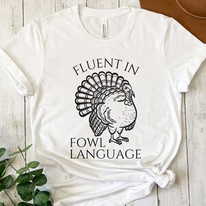 May include: White t-shirt featuring a black turkey illustration and the text "FLUENT IN FOWL LANGUAGE". The turkey is drawn with detailed feathers and a proud pose. The shirt is made of a soft material.