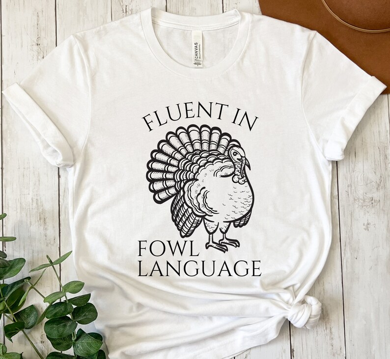 May include: White t-shirt featuring a black turkey illustration and the text "FLUENT IN FOWL LANGUAGE". The turkey is drawn with detailed feathers and a proud pose. The shirt is made of a soft material.
