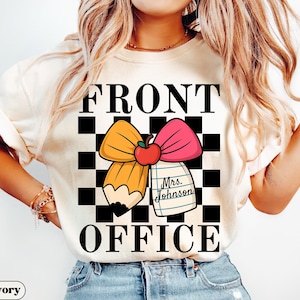 May include: Ivory-colored t-shirt with the words "FRONT OFFICE" in large black letters. The design includes a yellow pencil and pink bow with a red apple, set against a black and white checkered background. A tag reads "Mrs. Johnson".
