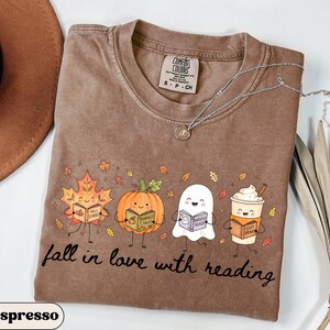 Peut inclure: Un t-shirt marron &agrave; manches courtes avec une impression graphique repr&eacute;sentant une feuille d'&eacute;rable, une citrouille, un fant&ocirc;me et une tasse de caf&eacute; souriants, tenant chacun un livre. Le texte indique fall in love with reading.