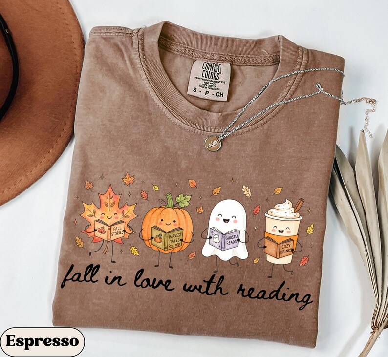 Peut inclure: Un t-shirt marron &agrave; manches courtes avec une impression graphique repr&eacute;sentant une feuille d'&eacute;rable, une citrouille, un fant&ocirc;me et une tasse de caf&eacute; souriants, tenant chacun un livre. Le texte indique fall in love with reading.