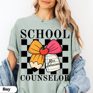 May include: A light green t-shirt with the words "SCHOOL COUNSELOR" in black block letters. The shirt features a graphic of a pencil and pink bow with a red apple and a notepad that says "Mrs. Johnson".
