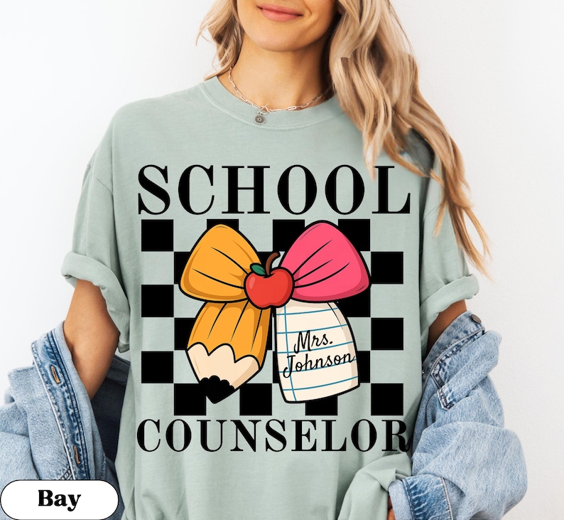 May include: A light green t-shirt with the words "SCHOOL COUNSELOR" in black block letters. The shirt features a graphic of a pencil and pink bow with a red apple and a notepad that says "Mrs. Johnson".