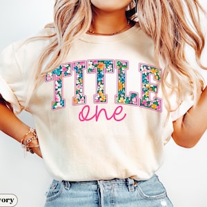 May include: Ivory-colored t-shirt with the words "TITLE one" in a floral design. The word "TITLE" is in large, block letters with a pink outline, filled with a colorful floral pattern. The word "one" is in a cursive, pink font.
