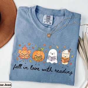 Peut inclure: Un t-shirt bleu jean avec un motif repr&eacute;sentant une feuille d'&eacute;rable, une citrouille, un fant&ocirc;me et une tasse de caf&eacute;, chacun tenant un livre. Le texte "fall in love with reading" est imprim&eacute; sous les personnages. &Eacute;tiquette Comfort Colors.