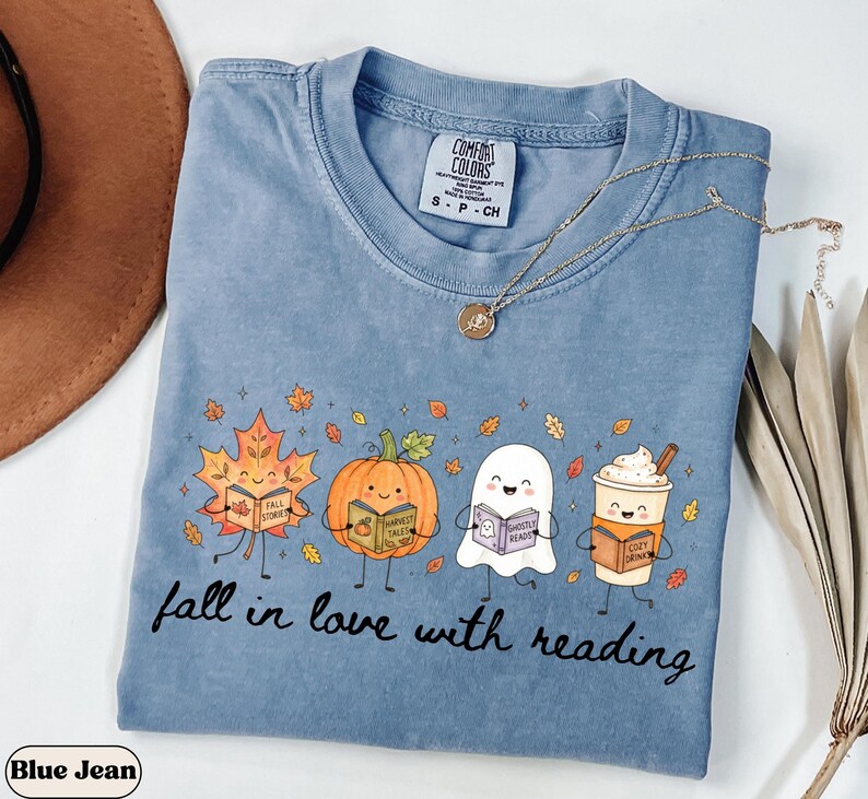 Peut inclure: Un t-shirt bleu jean avec un motif repr&eacute;sentant une feuille d'&eacute;rable, une citrouille, un fant&ocirc;me et une tasse de caf&eacute;, chacun tenant un livre. Le texte "fall in love with reading" est imprim&eacute; sous les personnages. &Eacute;tiquette Comfort Colors.