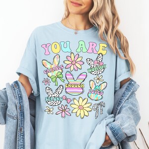 May include: Light blue t-shirt with the text "You Are" in rainbow colors. The shirt features a variety of colorful bunny and flower illustrations with positive affirmations like "Capable", "Worthy", "Enough", "Loved", and "Strong".